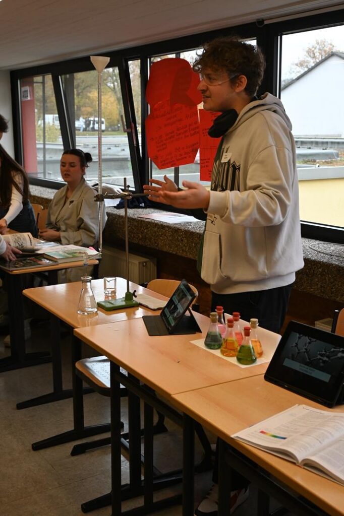 Schüler präsentiert ein Projekt in einem Klassenraum. Tische sind mit Materialien bedeckt.