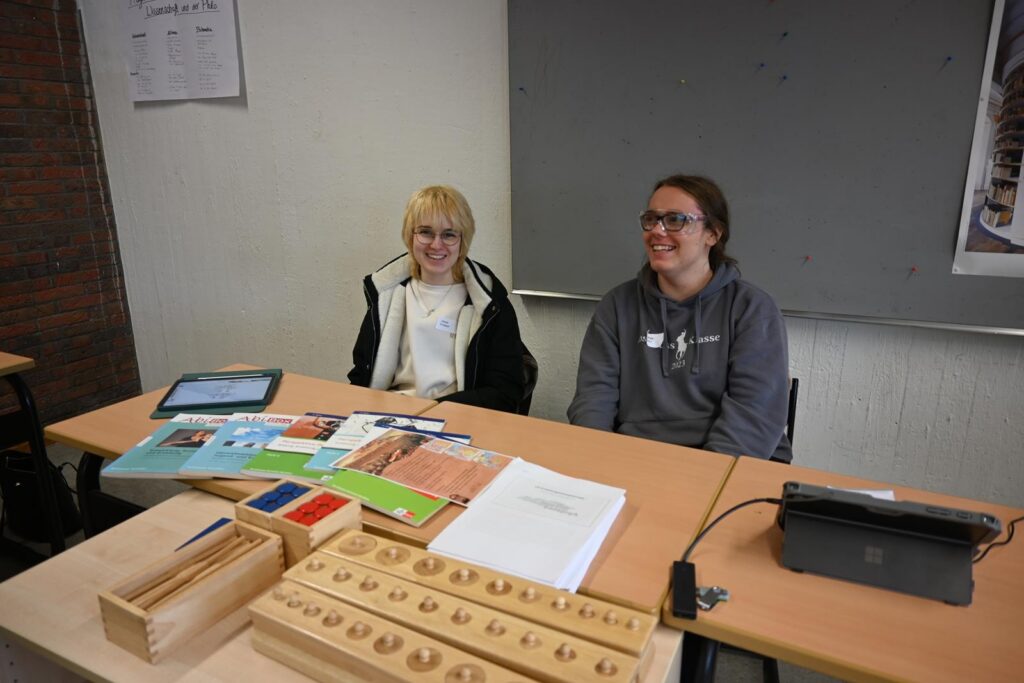 Zwei Personen sitzen an einem Tisch mit Unterrichtsmaterialien und einem Laptop.