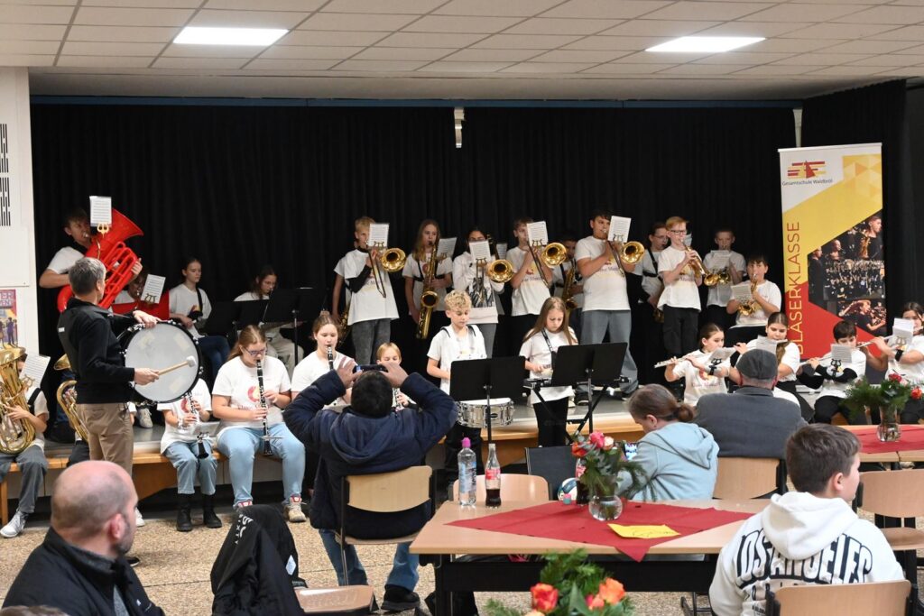 Schülerband spielt auf einer Bühne in einem Festsaal. Publikum hört zu.