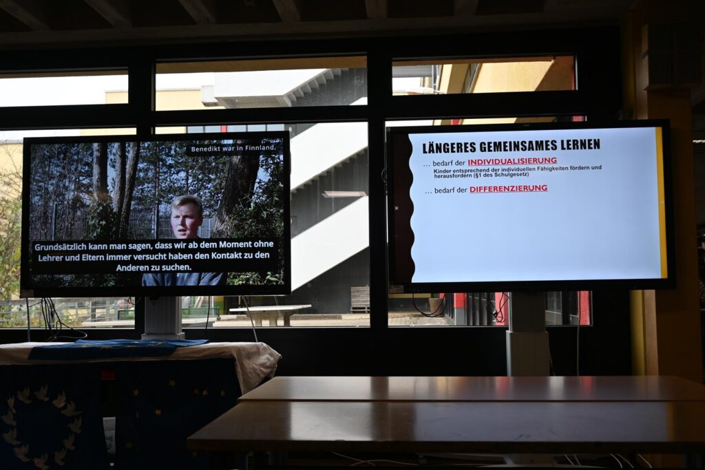 Zwei Monitore zeigen Informationen und einen Videoausschnitt in einem Klassenraum.
