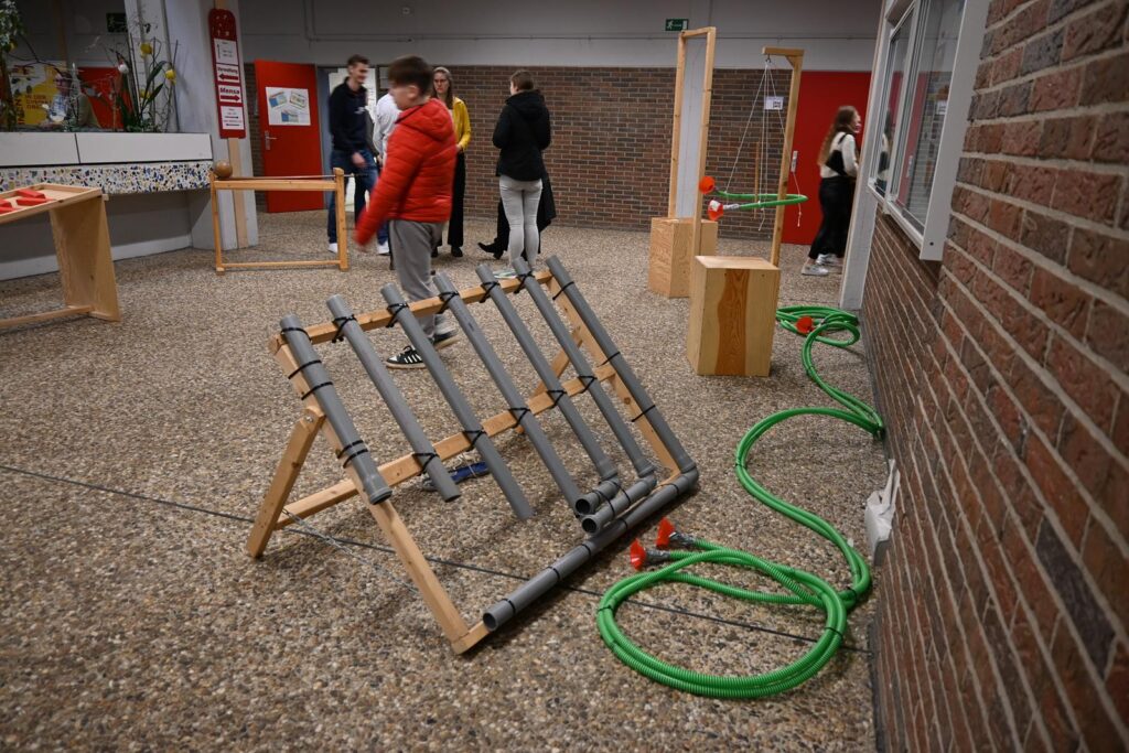 Holzgestell mit Rohren und Schläuchen in einem Raum, Schüler interagieren damit.