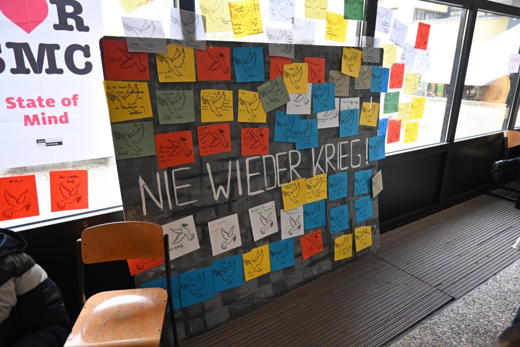 Bunte Zettel mit der Aufschrift "NIE WIEDER KRIEG" an einer Wand.