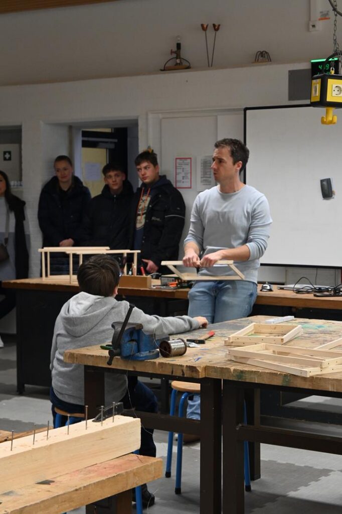 Lehrer erklärt Schülern ein Projekt in einer Werkstatt. Hinter ihnen stehen weitere Schüler.