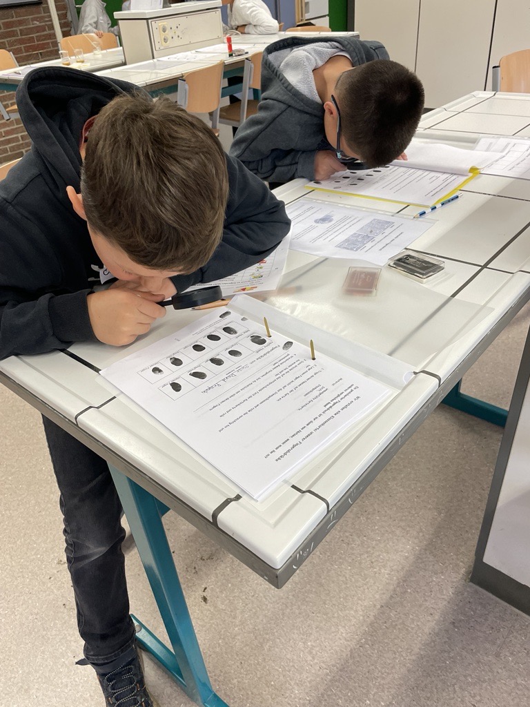 Zwei Schüler arbeiten an einem Tisch und betrachten Aufgabenblätter mit Lupe.