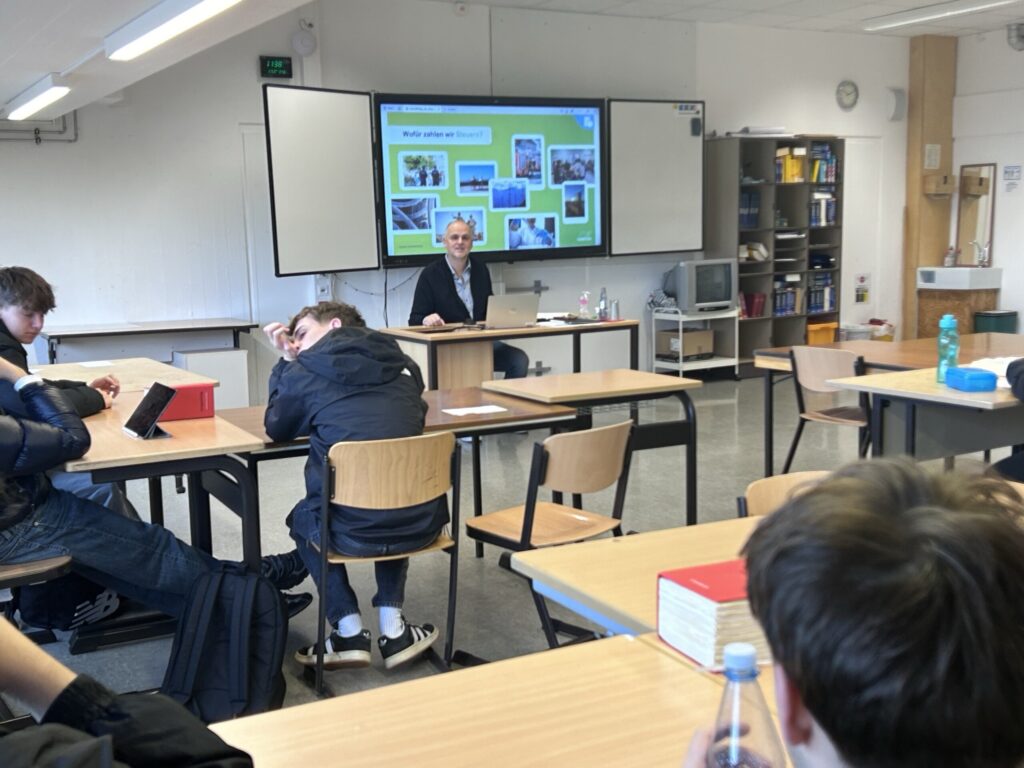 Ein Lehrer präsentiert Inhalte in einem Klassenzimmer. Schüler sitzen an Tischen.