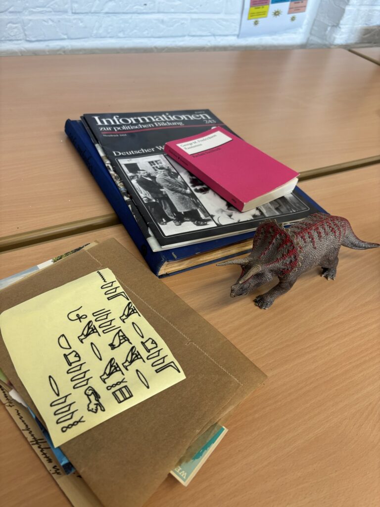 Auf einem Tisch liegen Bücher, Notizblätter und eine Dinosaurierfigur.