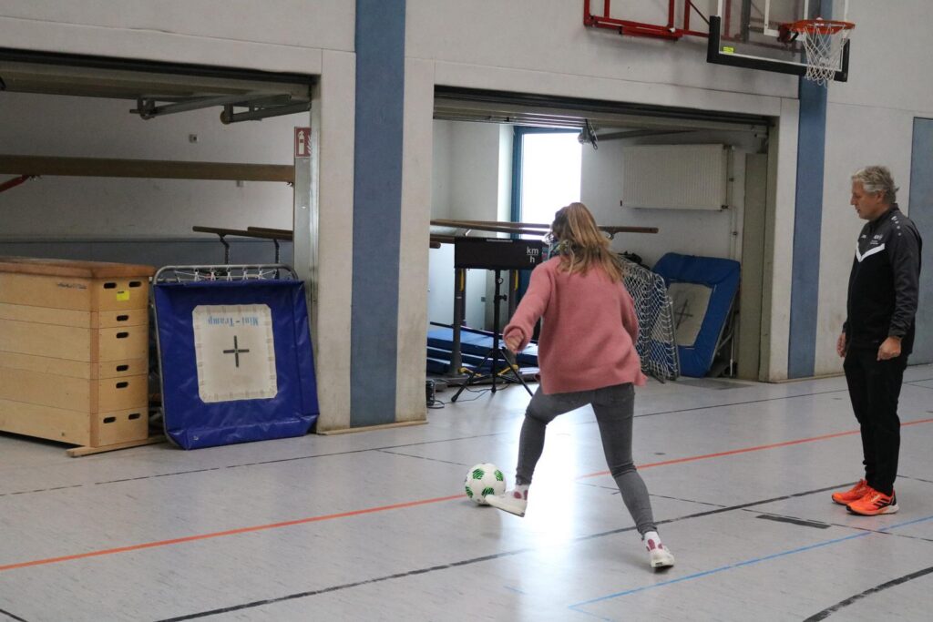 Mädchen spielt Fußball in einer Sporthalle, Trainer beobachtet.