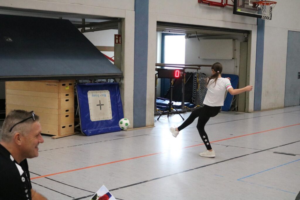 Ein Mädchen schießt einen Fußball in einer Sporthalle, ein Zuschauer sitzt daneben.