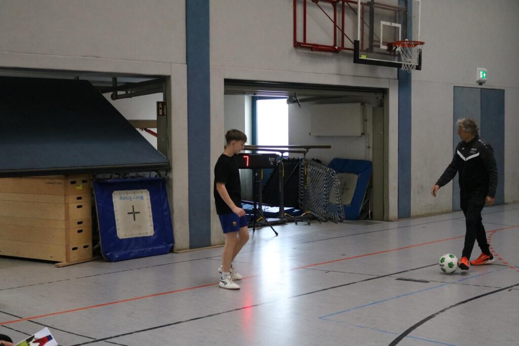 Junge spielt Fußball in einer Sporthalle mit Trainer.