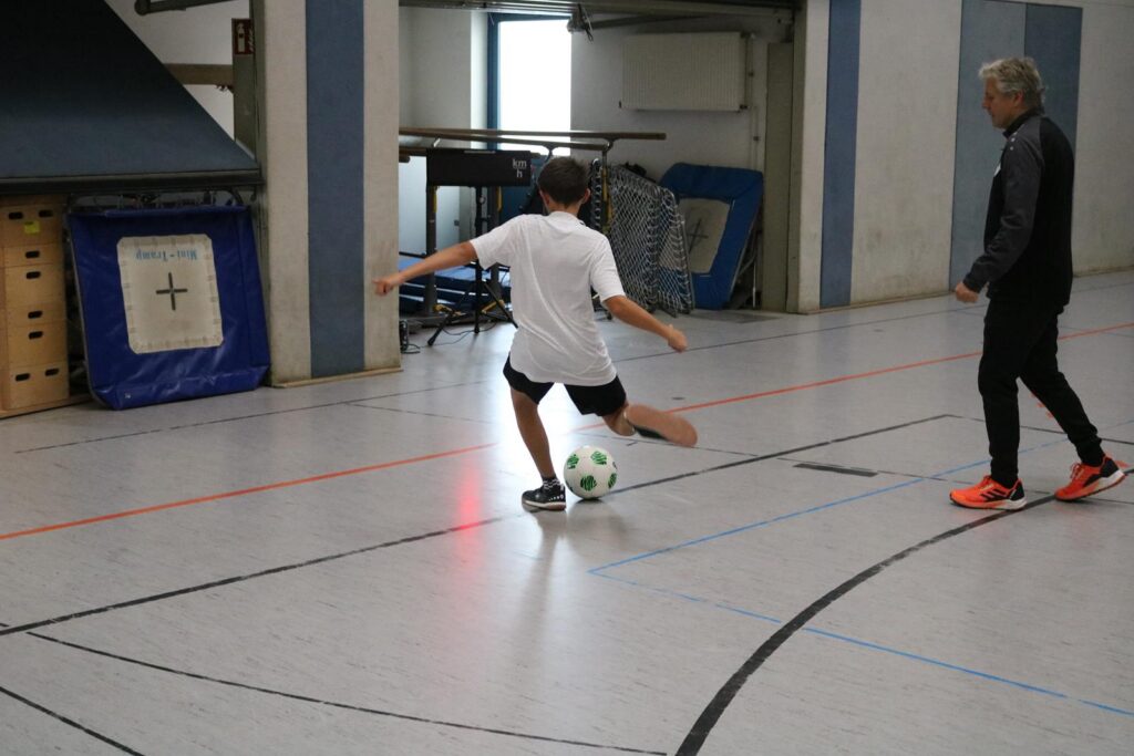 Ein Junge kickt einen Fußball in einer Sporthalle, ein Trainer beobachtet ihn.
