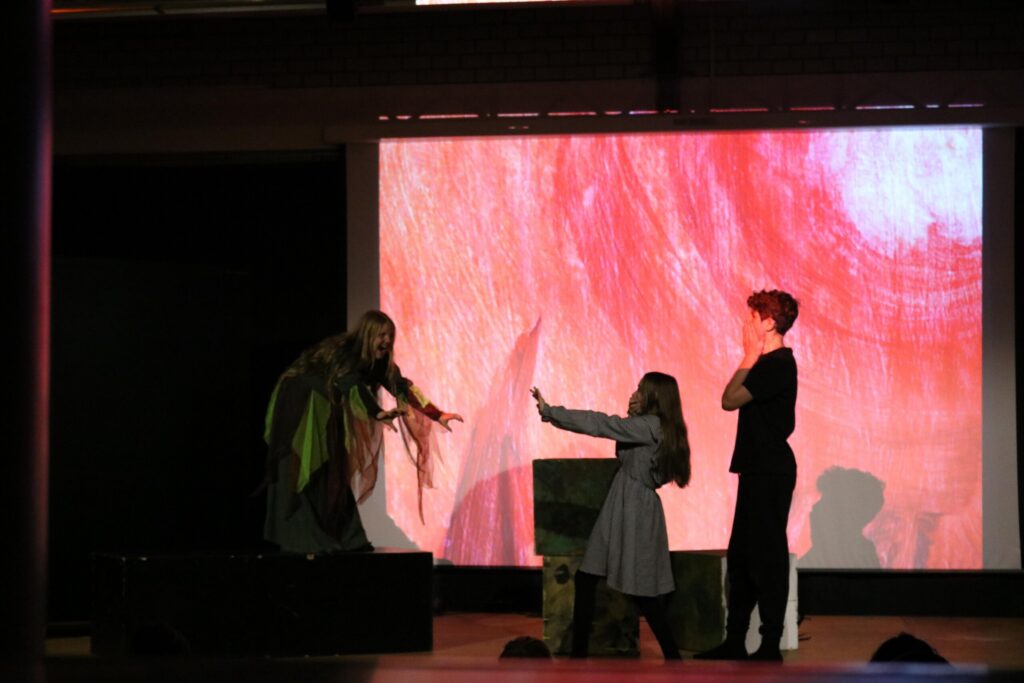 Theateraufführung mit schauspielenden Kindern auf der Bühne. Bühnenbild und Projektion im Hintergrund.