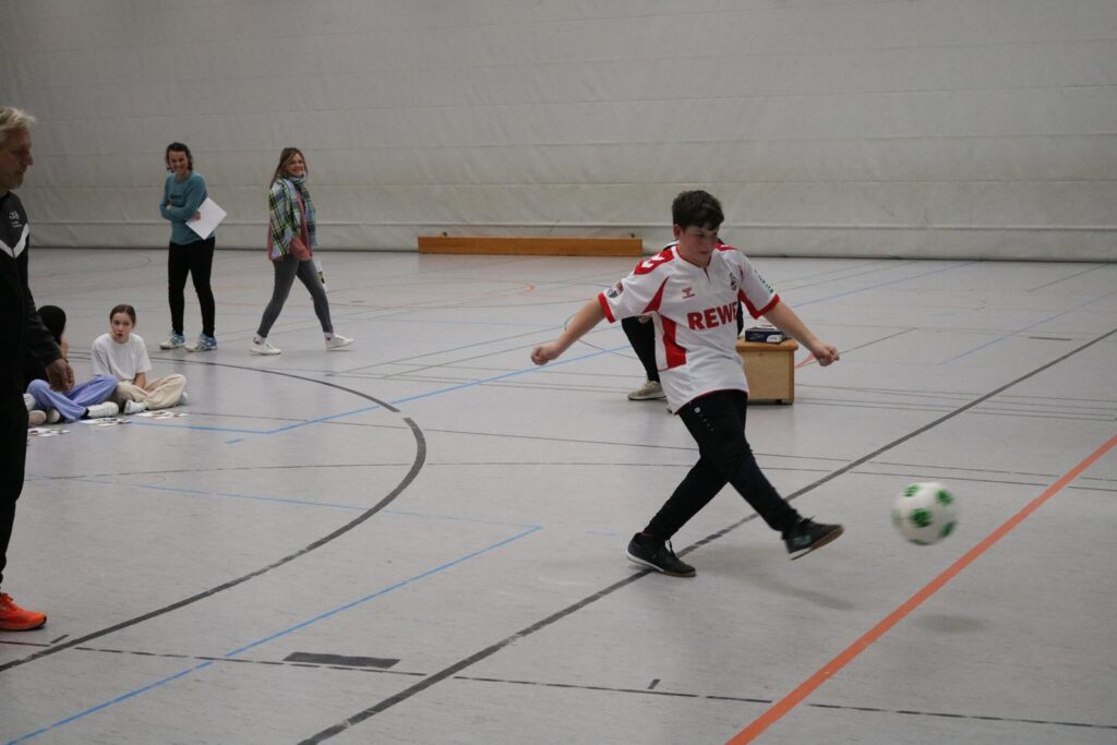 Ein Junge kickt einen Fußball in einer Sporthalle, Zuschauer sitzen im Hintergrund.