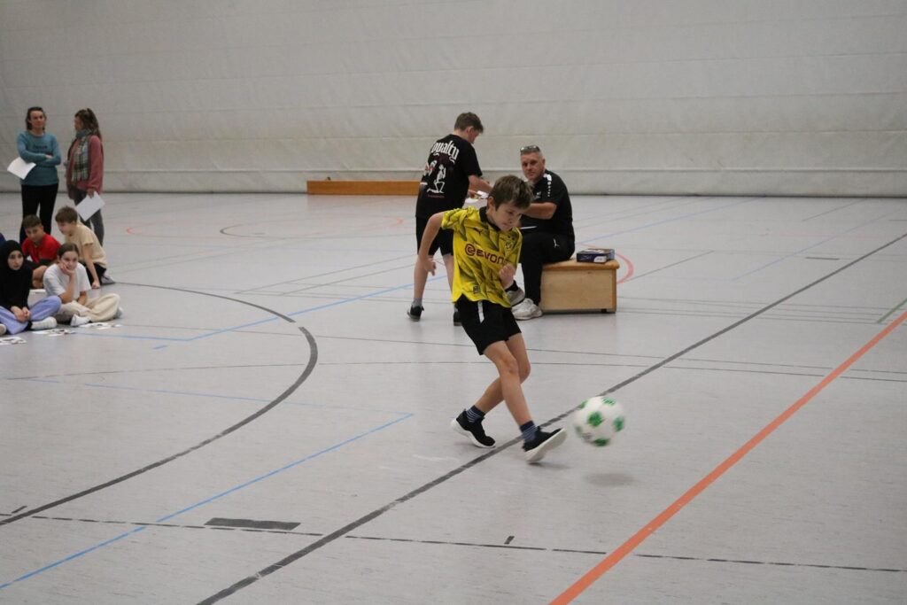 Junge in gelber Kleidung spielt Fußball in einer Sporthalle.
