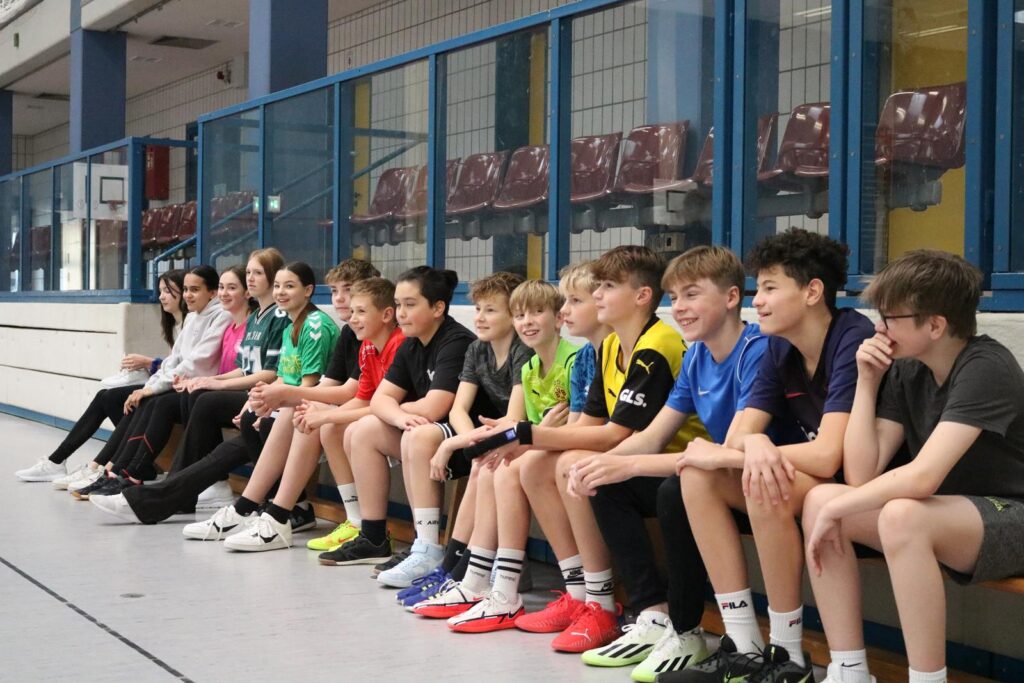 Schülerinnen und Schüler sitzen in einer Sporthalle auf einer Bank.