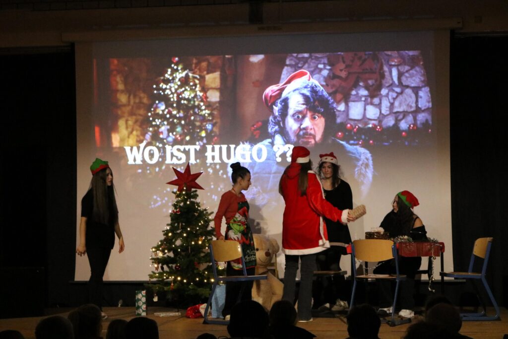 Theateraufführung mit Weihnachtsbaum, Schauspielern in Kostümen und Projektion im Hintergrund.