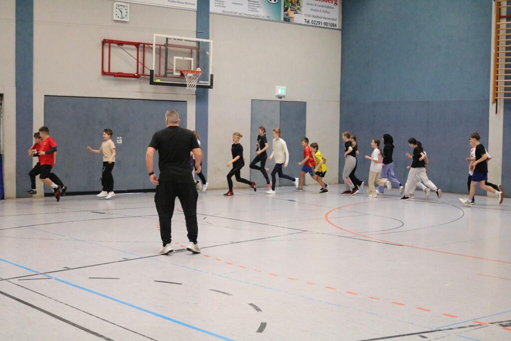 Schülerinnen und Schüler joggen in einer Turnhalle. Ein Lehrer steht vorne.
