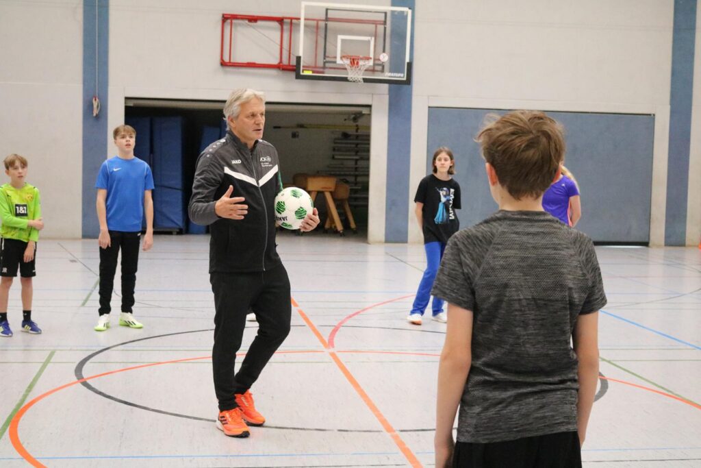 Trainer erklärt eine Übung mit Kindern in einer Sporthalle.