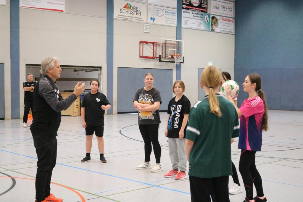 Ein Trainer erklärt einer Gruppe von Sportlerinnen in der Halle eine Übung.