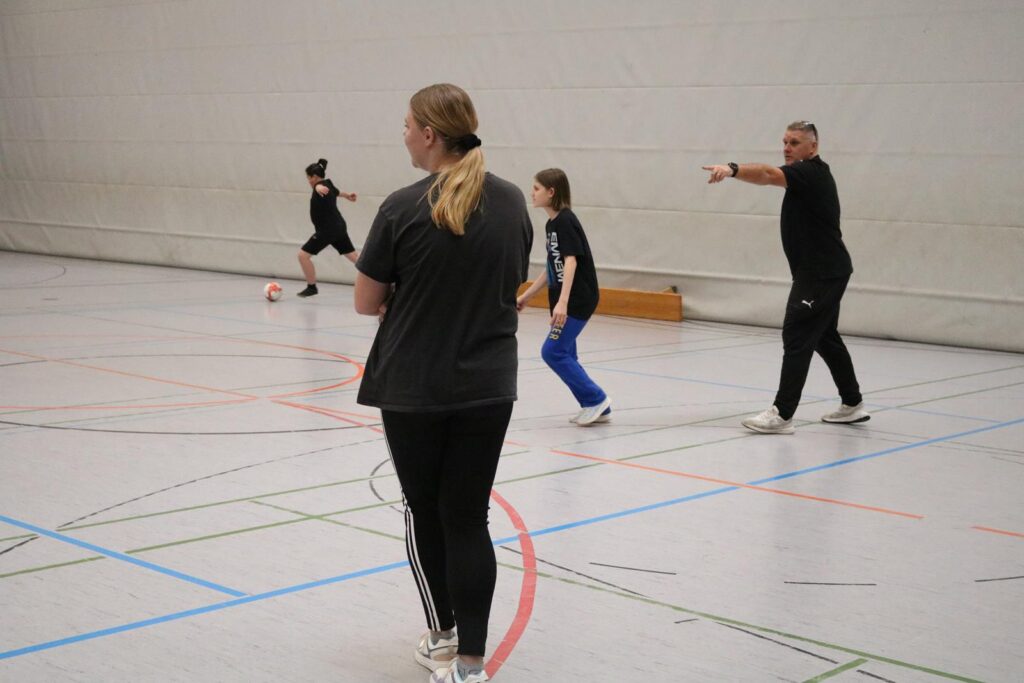 Sporthalle mit mehreren Personen beim Training, eine Person gibt Anweisungen.