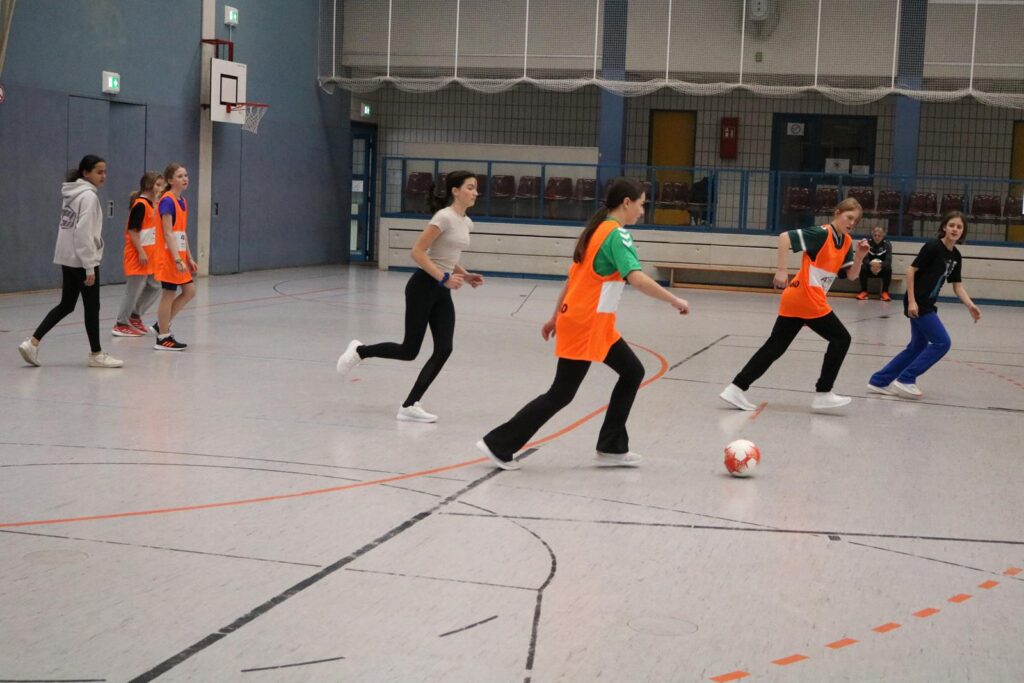 Mädchen spielen Fußball in einer Sporthalle. Einige tragen orangefarbene Westen.