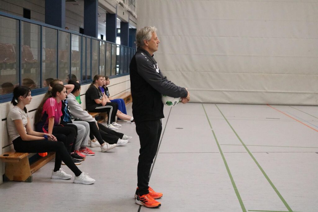 Ein Trainer steht in einer Sporthalle, während Schüler auf Bänken sitzen.