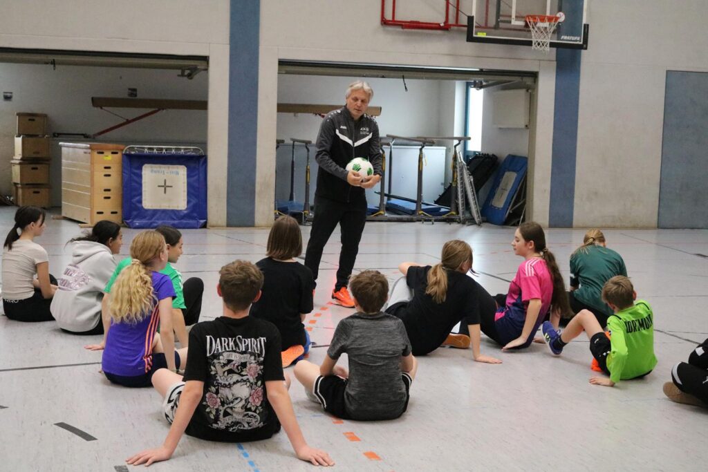 Trainer spricht mit Kindern in einer Sporthalle, Fußball in der Hand.