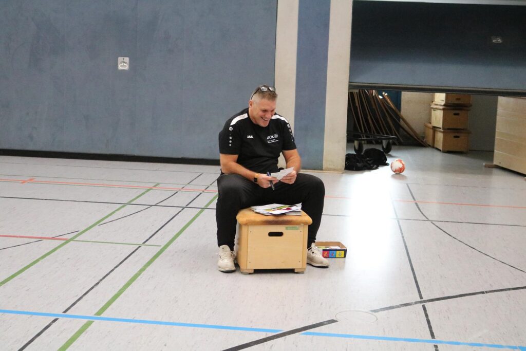 Ein Mann sitzt auf einem Kasten in einer Sporthalle und liest Unterlagen.