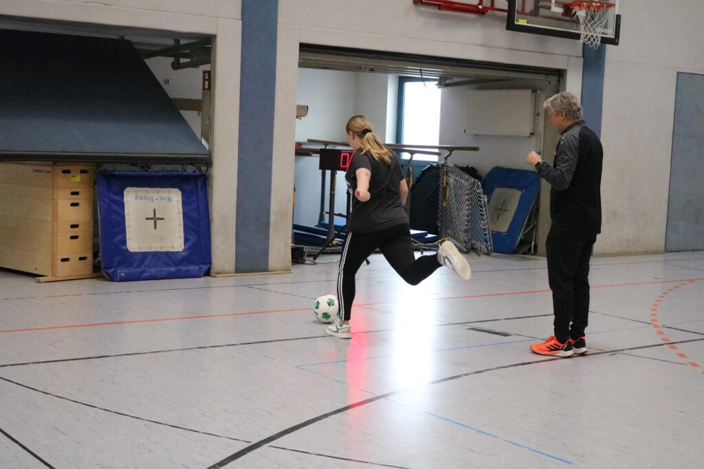 Eine Person dribbelt einen Fußball in einer Sporthalle, ein Trainer beobachtet.