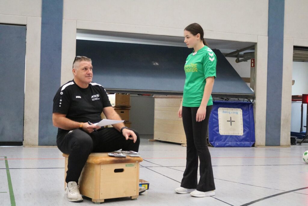 Trainer sitzt auf einer Kiste, während eine Spielerin im grünen Shirt zuhört.