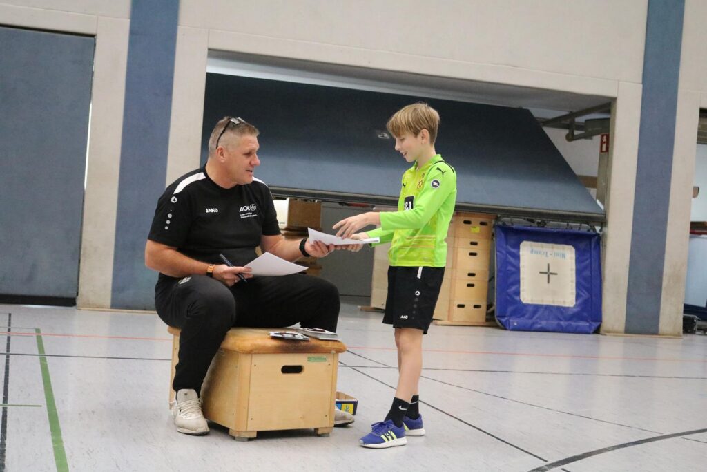 Ein Trainer sitzt auf einer Kiste und spricht mit einem Jungen in Sportkleidung.