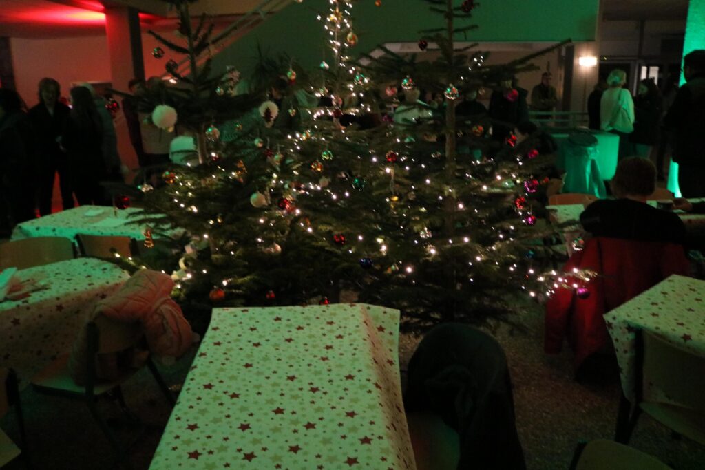 Weihnachtsbaum mit Lichtern und Dekoration in einem festlichen Raum. Tische mit roten Tischdecken.