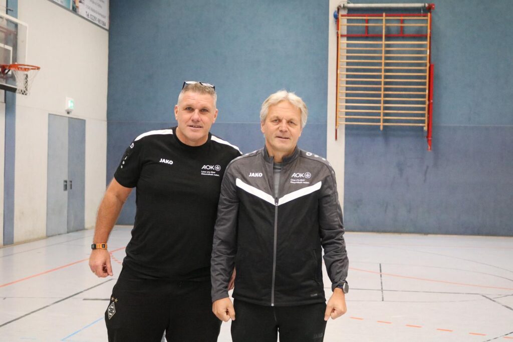 Zwei Männer in Sportbekleidung stehen in einer Turnhalle.