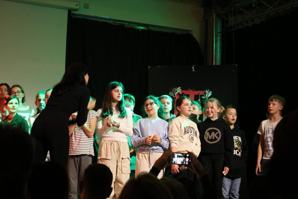 Schüler stehen auf der Bühne während einer Aufführung mit einem Publikum im Hintergrund.