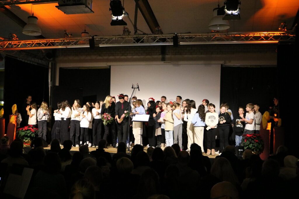 Gruppenvorstellung auf der Bühne mit vielen Teilnehmern und Publikum im Hintergrund.