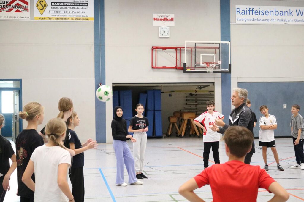 Sporthalle mit Schülern, die an einer Sportstunde teilnehmen. Ein Lehrer erklärt eine Übung.
