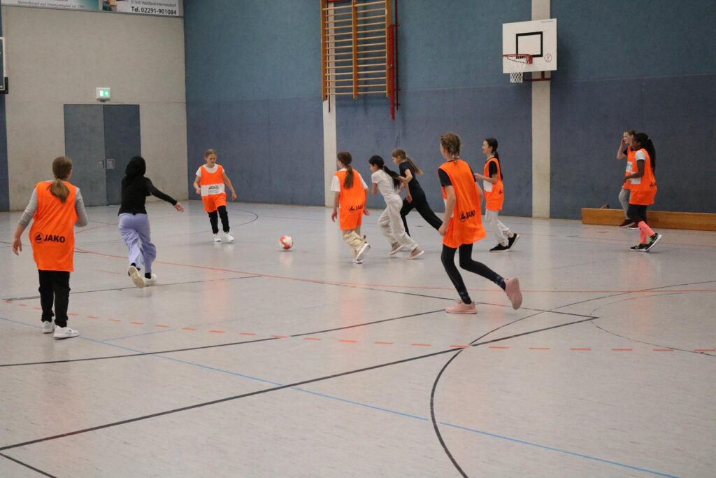 Mädchen spielen Fußball in einer Sporthalle. Einige tragen orangefarbene Westen.