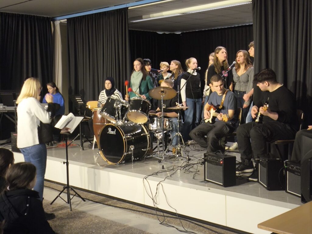 Schülerband spielt auf der Bühne, Sänger und Instrumentalisten sind aktiv.