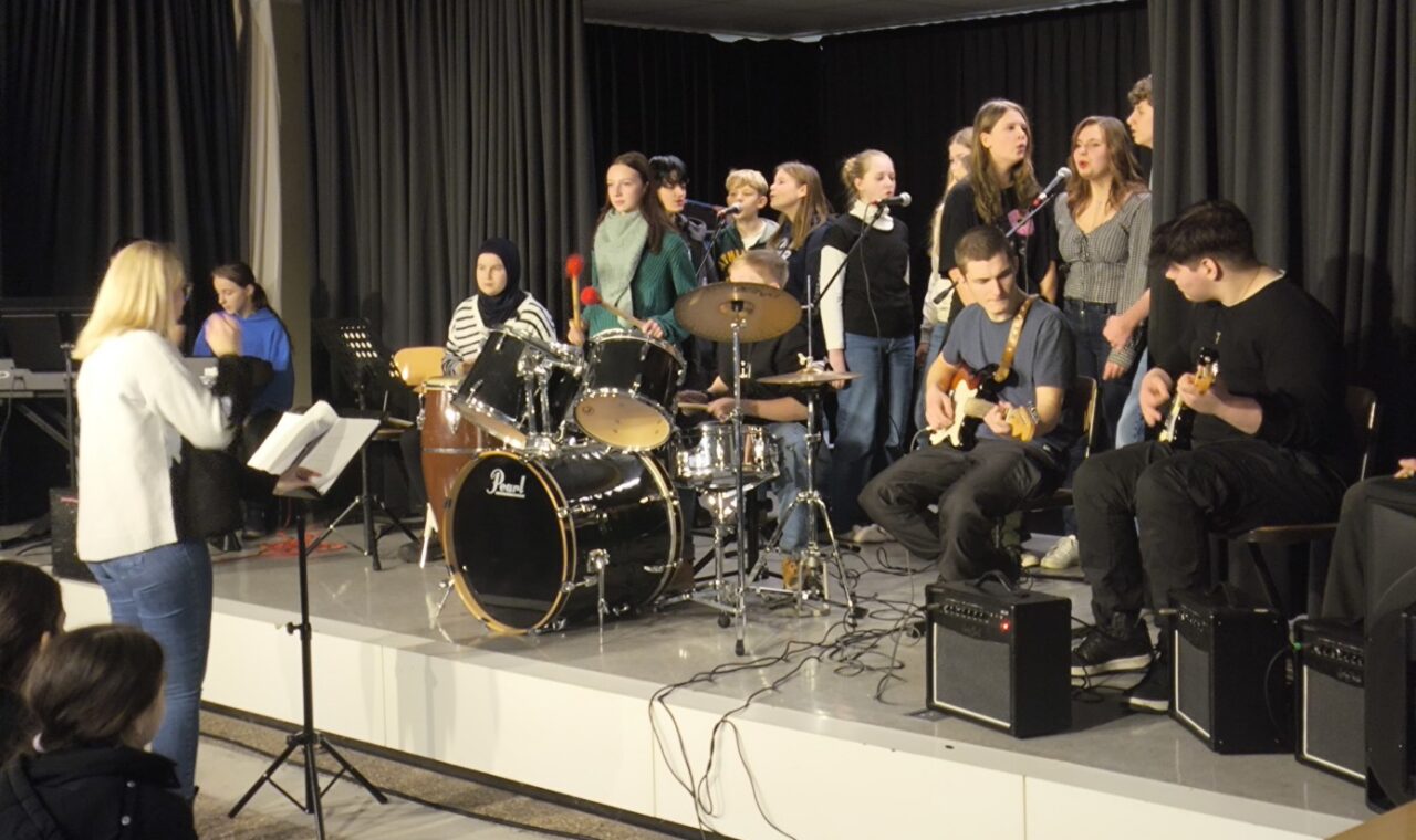 Schülerband spielt auf der Bühne, Sänger und Instrumentalisten sind aktiv.