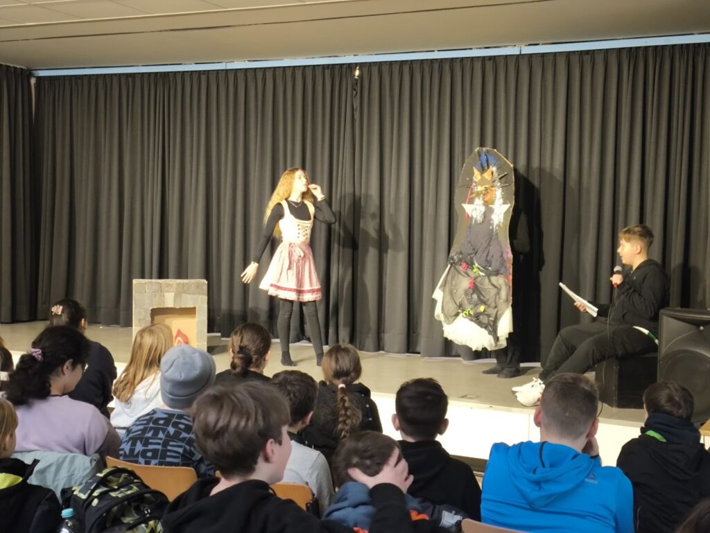Schüler führt auf einer Bühne ein Theaterstück vor, Publikum sitzt davor.