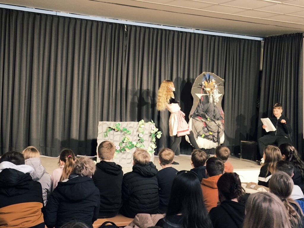 Schüler führen ein Theaterstück auf einer Bühne vor. Zuschauer sitzen im Publikum.