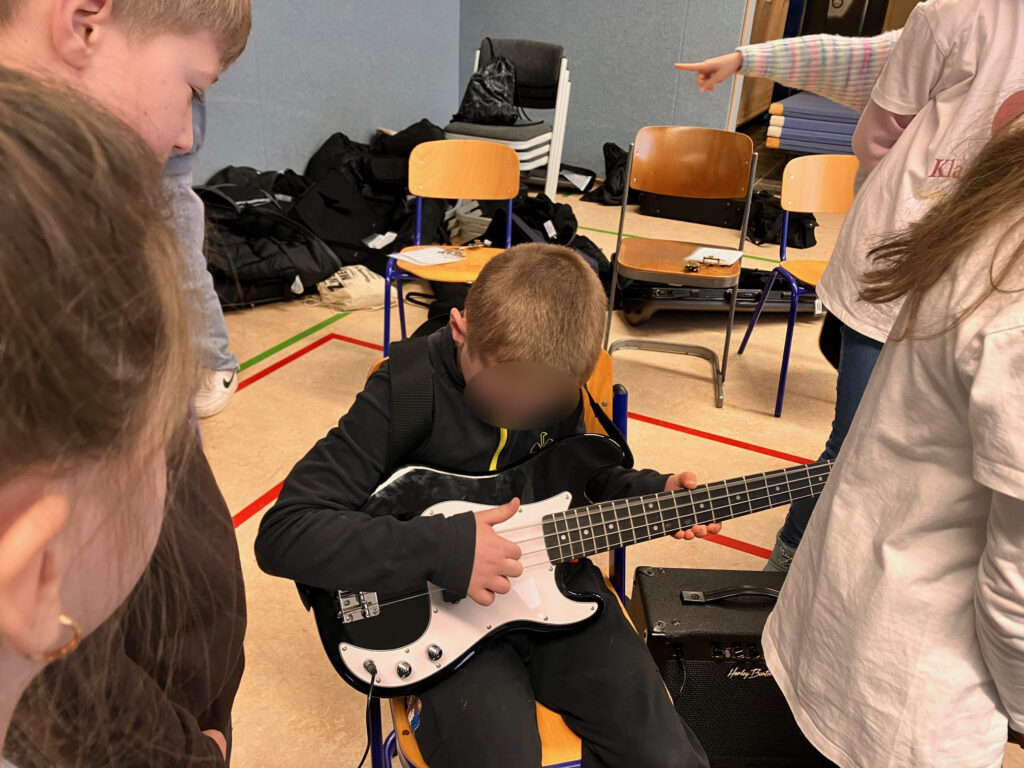 Ein Kind spielt E-Gitarre, umgeben von anderen Kindern. Stühle im Hintergrund.