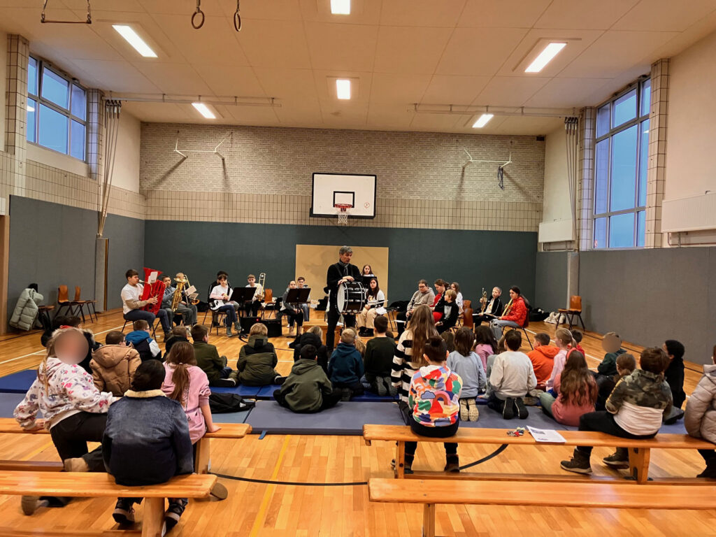 Schüler sitzen auf Bänken in einer Turnhalle, während ein Ensemble spielt.