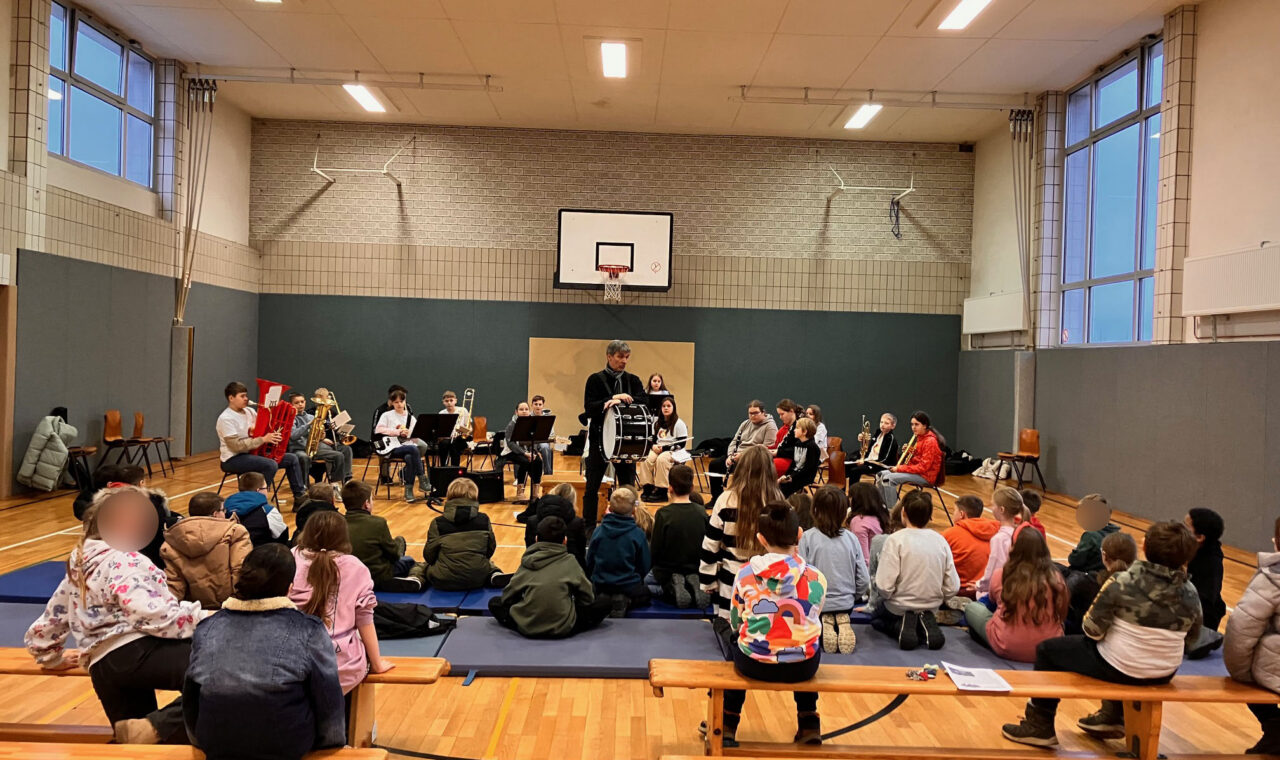 Schüler sitzen auf Bänken in einer Turnhalle, während ein Ensemble spielt.