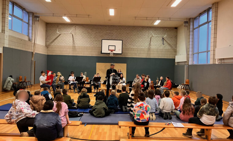 Schüler sitzen auf Bänken in einer Turnhalle, während ein Ensemble spielt.