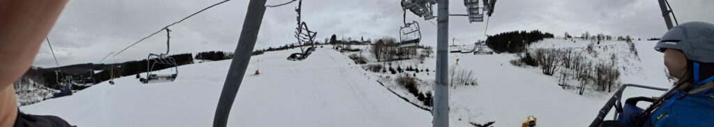 Personen auf einem Skilift mit verschneiter Landschaft im Hintergrund.