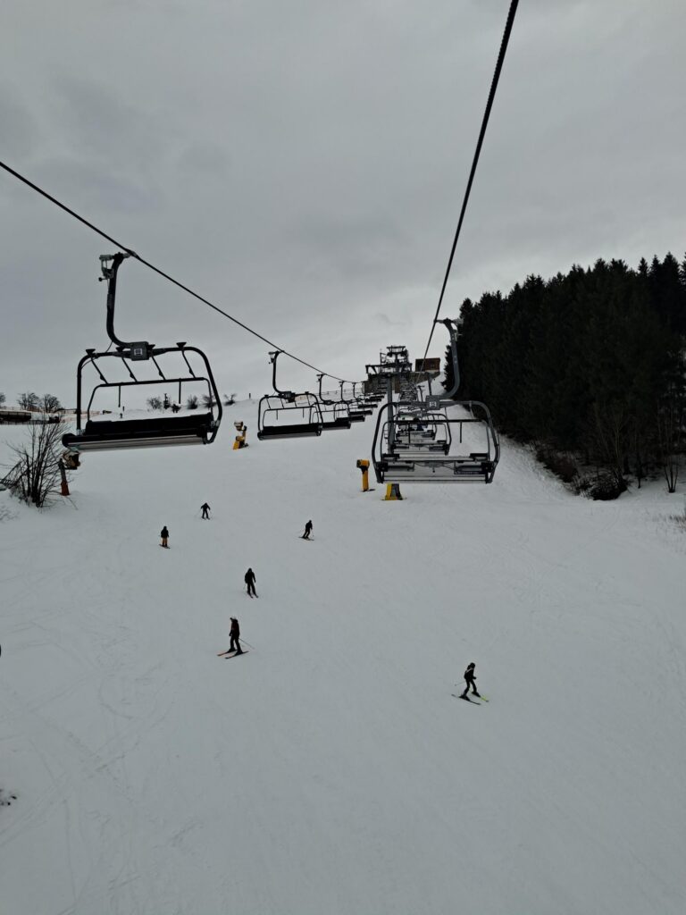 Skigebiet mit Sessellift und Skifahrern auf der Piste bei bewölktem Himmel.