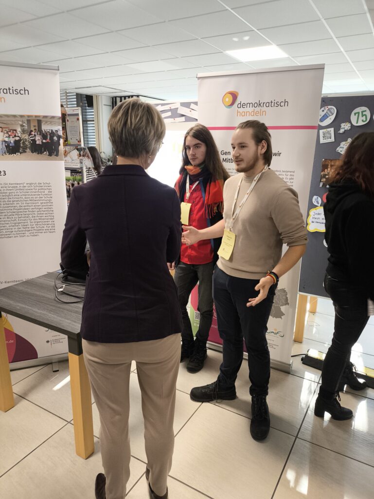 Drei Personen stehen an einem Informationsstand in einem hellen Raum.
