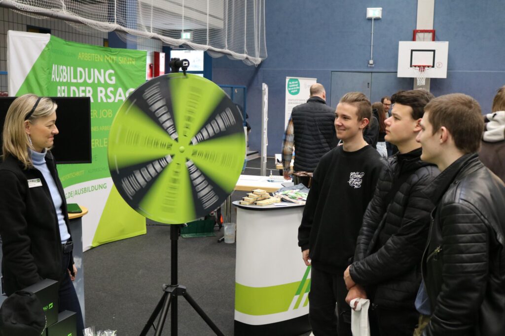 Messebesucher stehen vor einem Stand und sehen sich ein Gewinnrad an.