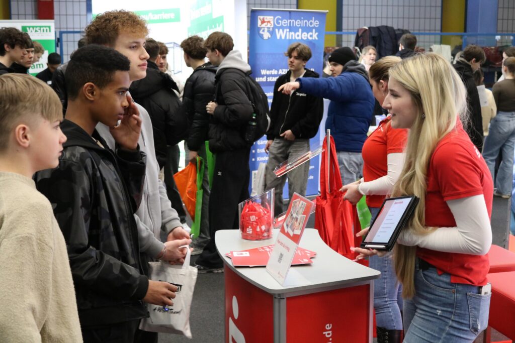 Schüler stehen an einem Stand, während eine Mitarbeiterin Informationen gibt.