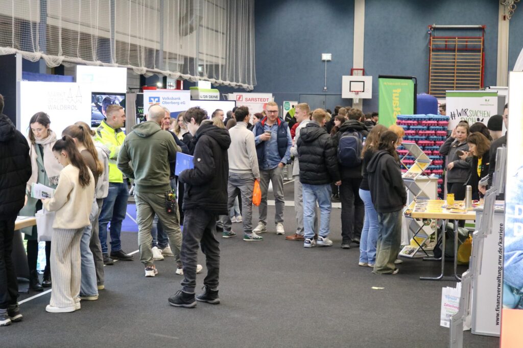 Schüler und Besucher auf einer Messe in einer Sporthalle, zahlreiche Stände.